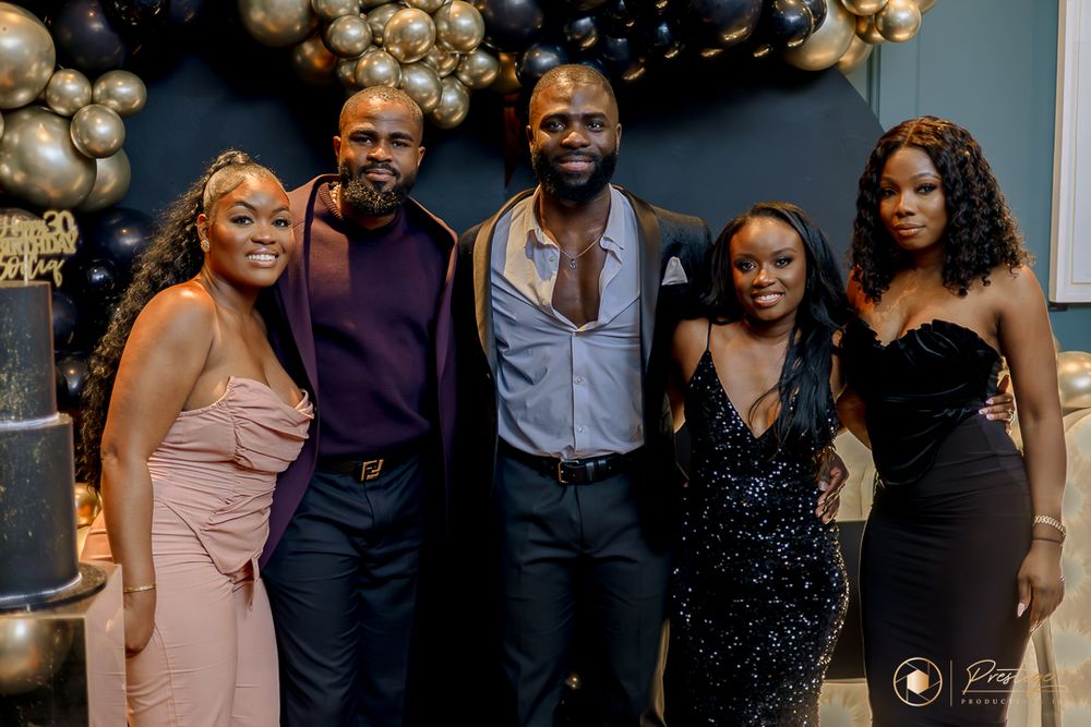 Glamorous celebration with partygoers in formal attire posing against black and gold balloon backdrop.