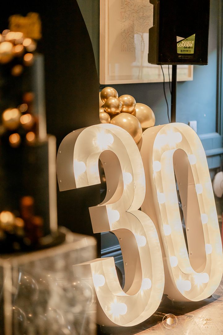 Illuminated number 30 marquee sign with gold balloon decorations at an elegant party.