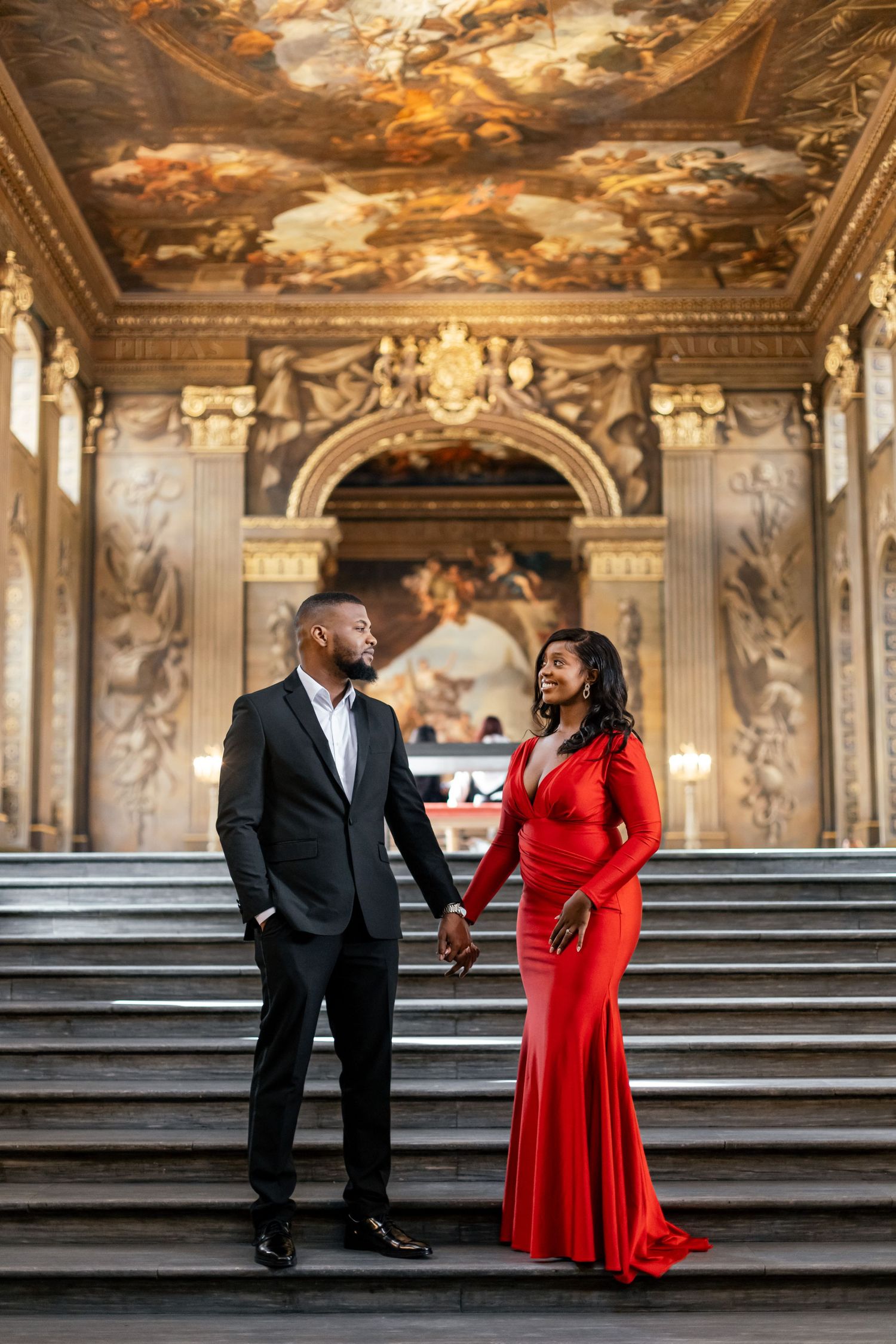 Elegant couple posing on grand staircase with ornate painted ceiling and classical architecture.