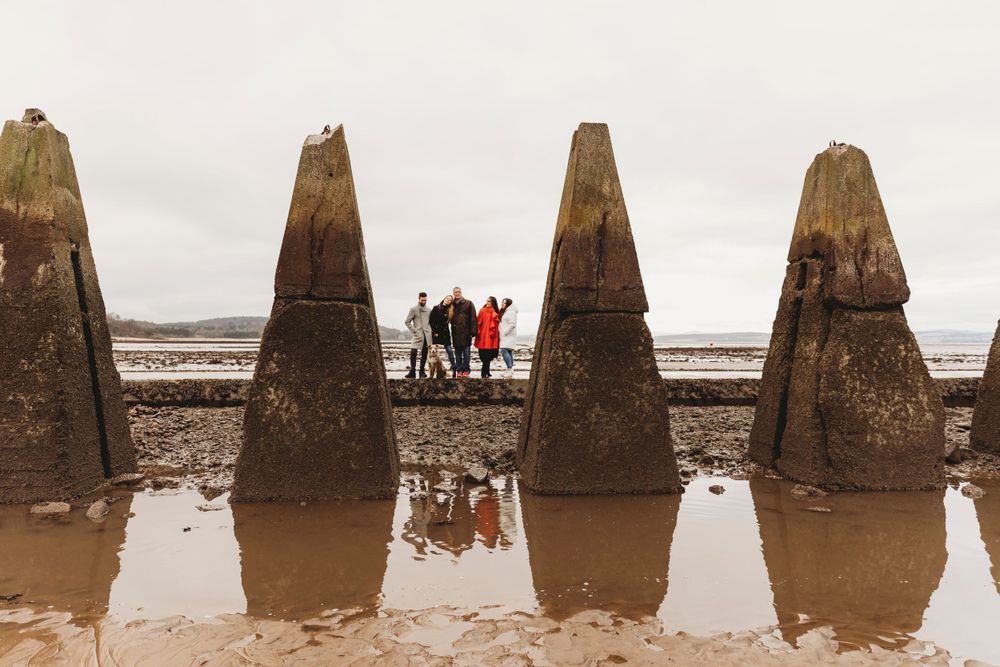 Four tall concrete pillars stand on a beach with silhouettes of people reflected in water puddles at their base.