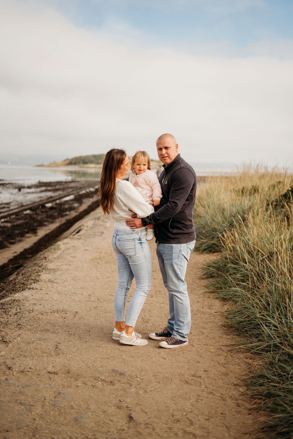 Family enjoying a tender moment together on a coastal path with beach grass and ocean views in the background.