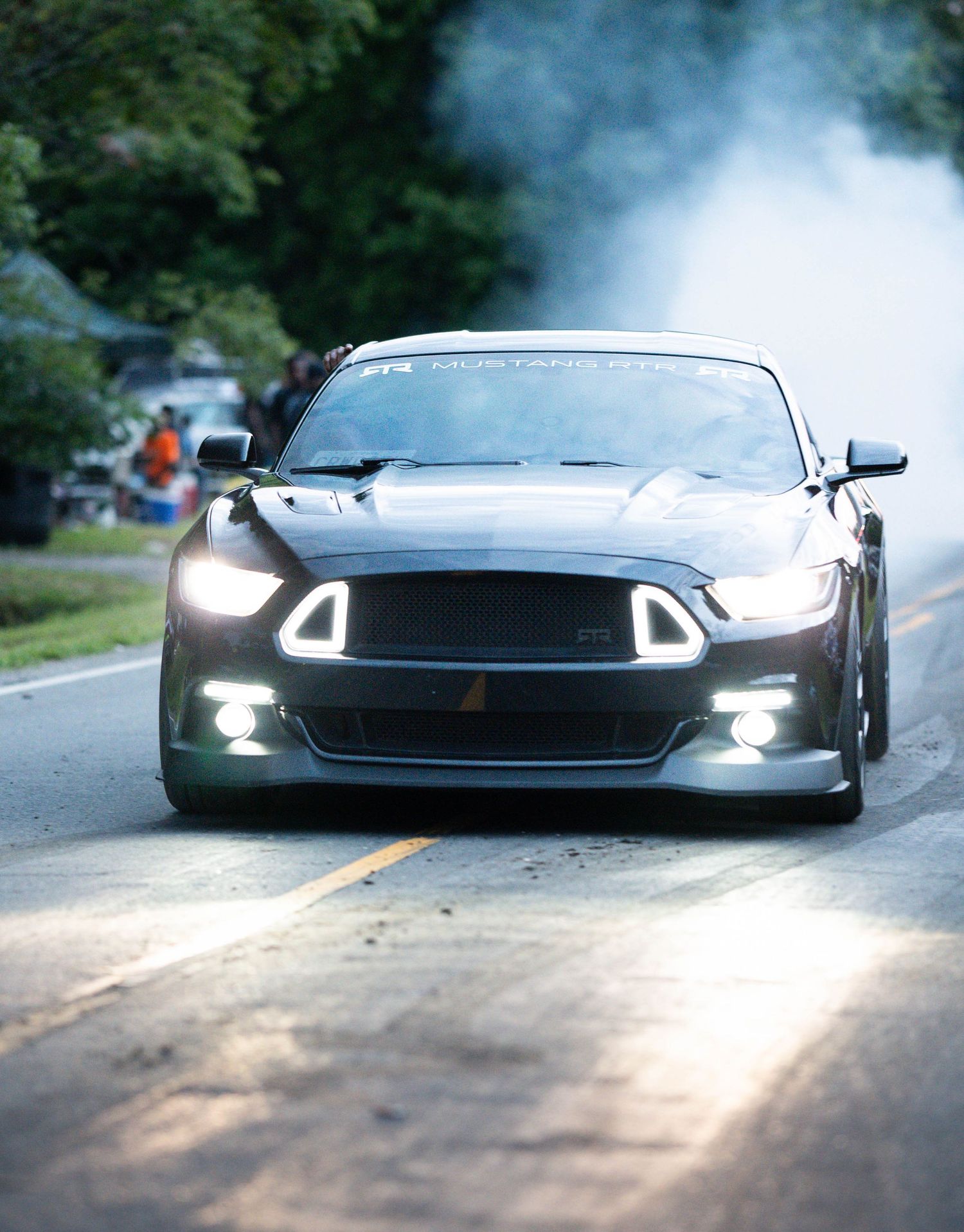 Black Ford Mustang with illuminated LED headlights driving on a misty forest road.