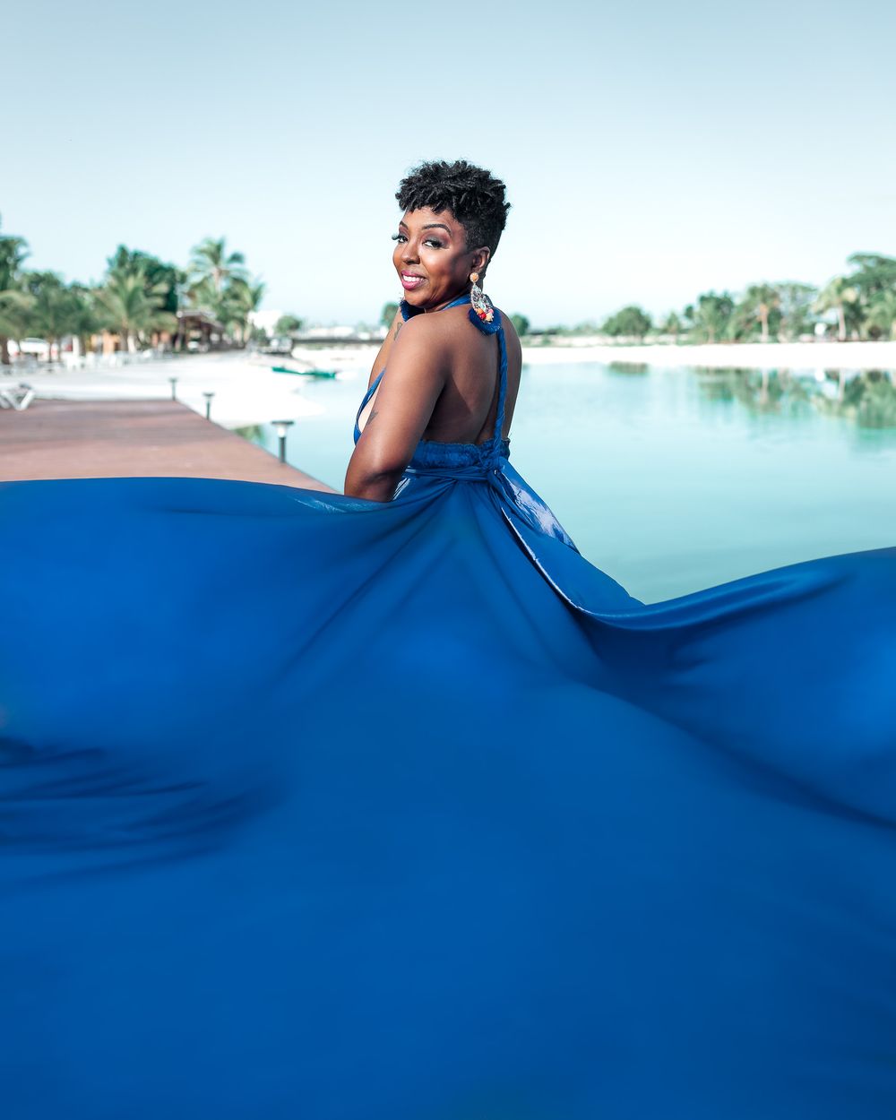 A flowing royal blue dress billows dramatically against a tranquil waterfront backdrop.