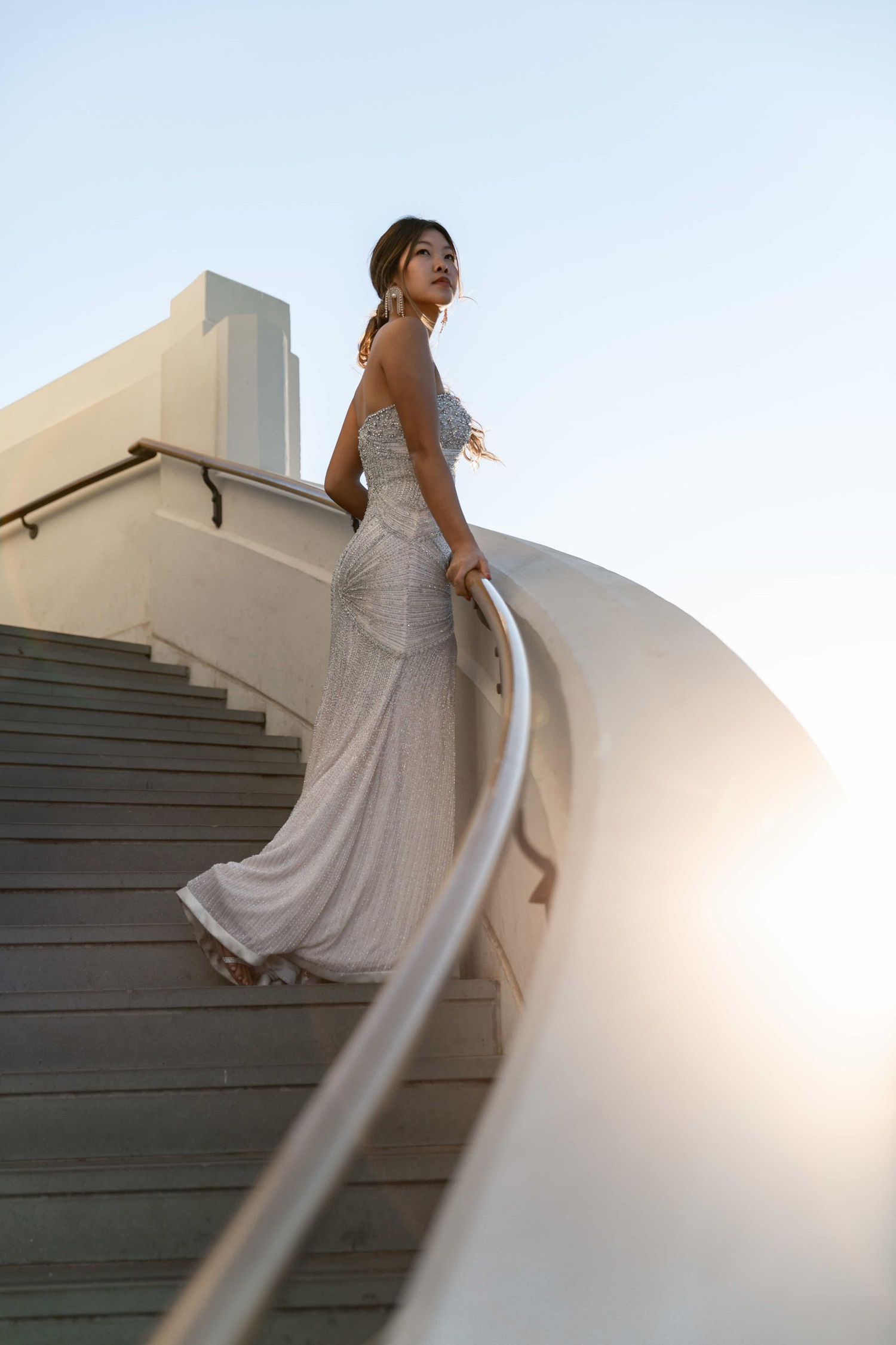 Bride in silver beaded gown stands on curved staircase with dramatic sunset backlight behind her.