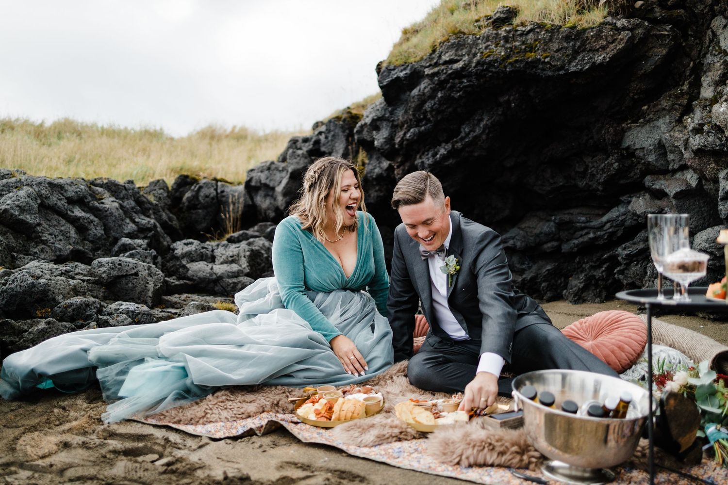 Romantic coastal picnic setup with champagne and charcuterie board on rocks by the ocean.