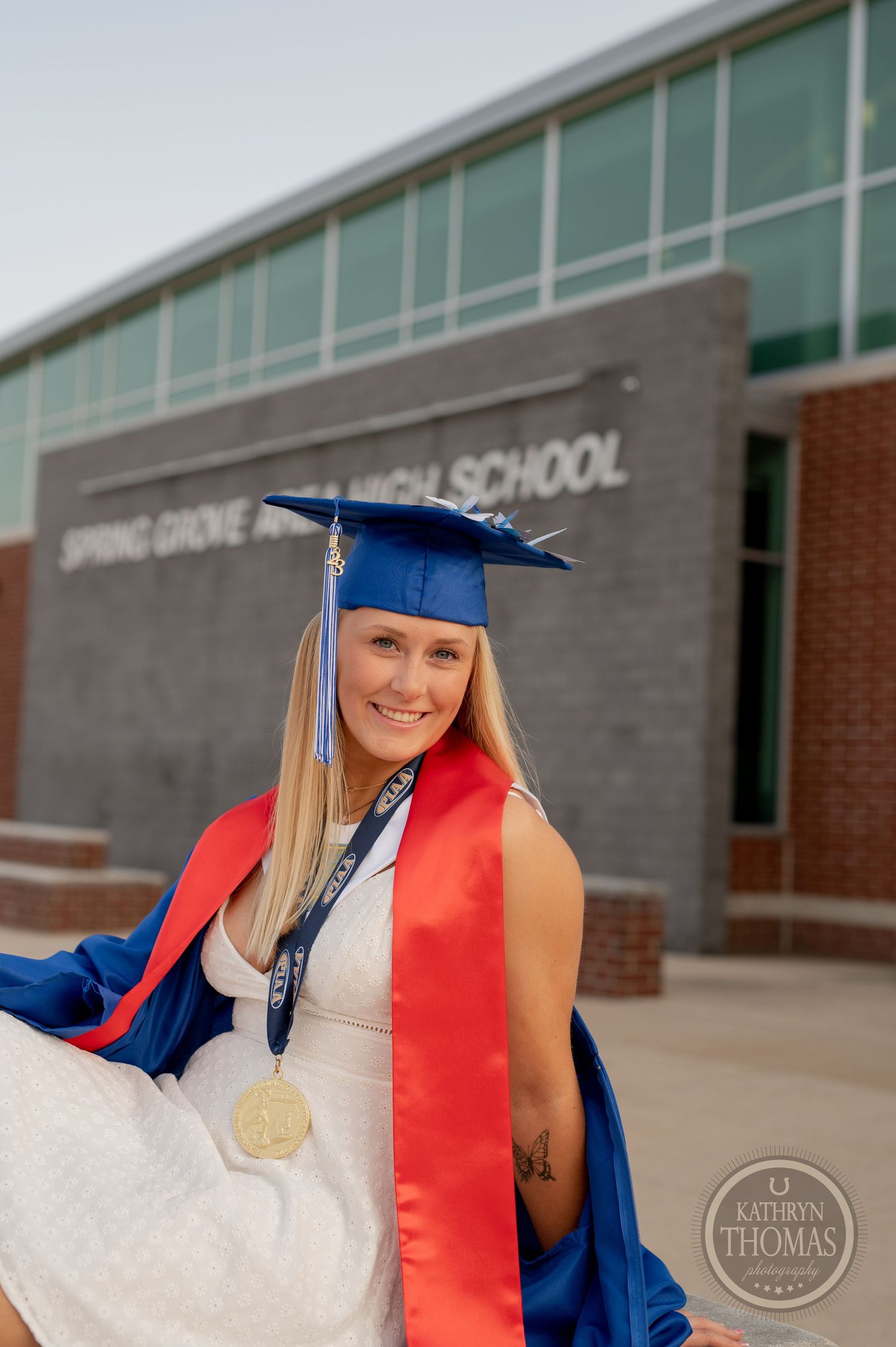 Graduate in cap and gown with red stole smiles in front of Spring Grove Area High School.