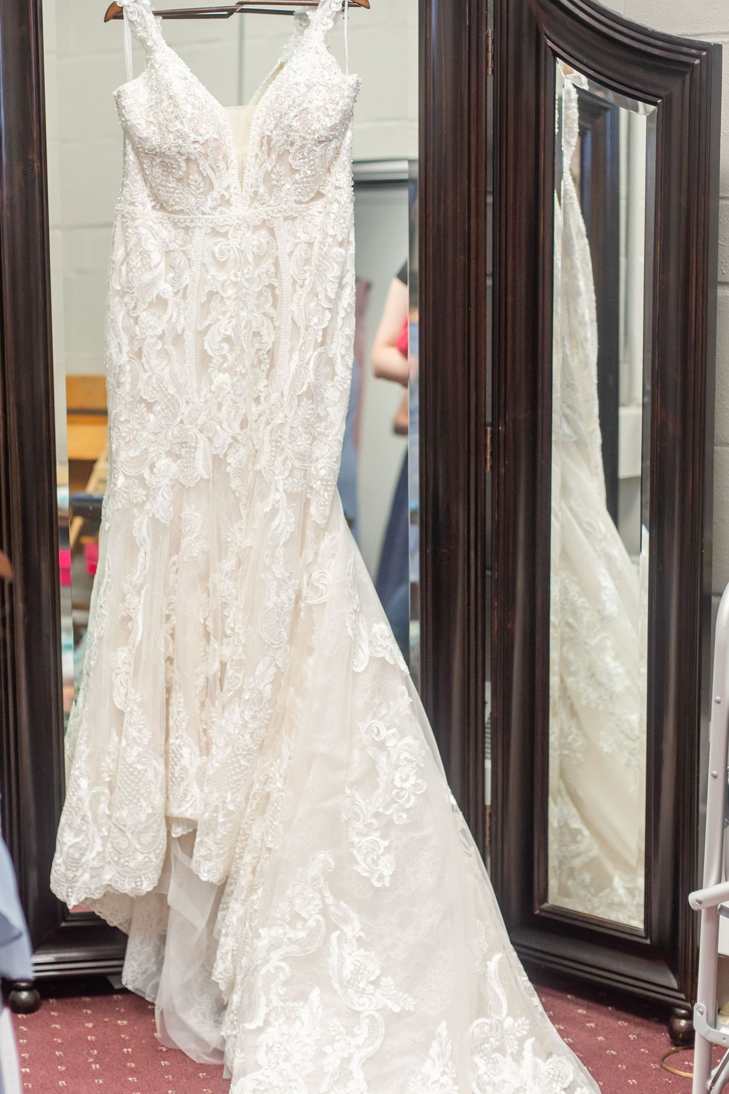 White lace mermaid wedding gown with intricate embroidery displayed in front of ornate mirrors.