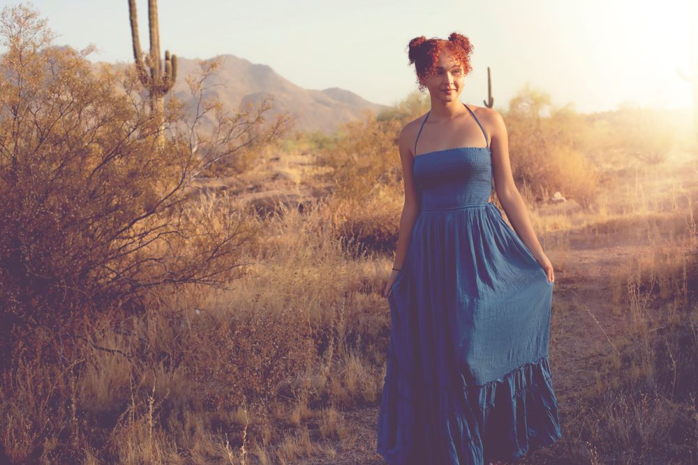 Figure in flowing blue dress stands among desert plants at sunset.