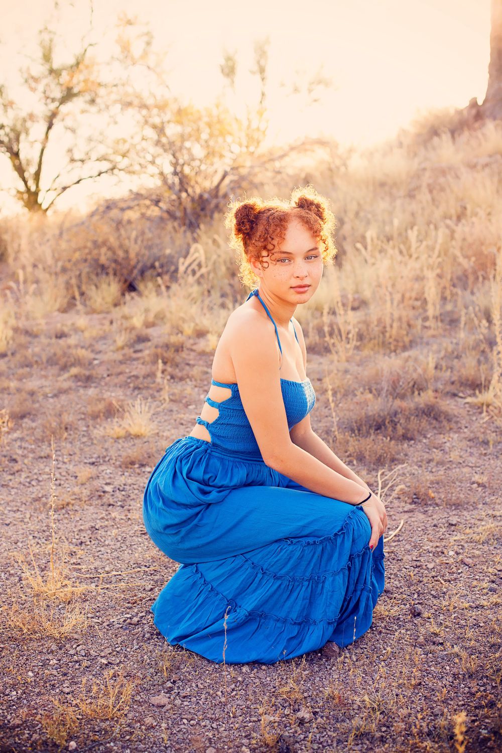 A person in a vibrant blue dress crouches in a desert landscape during golden hour.