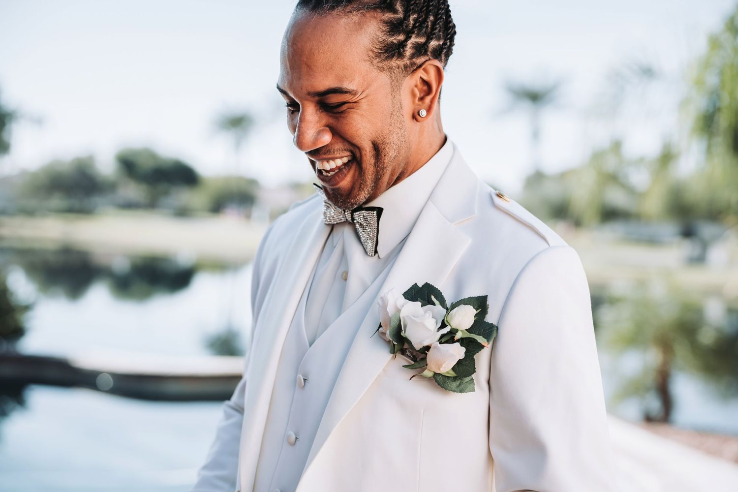 Groom in white tuxedo with boutonniere smiling by waterfront.