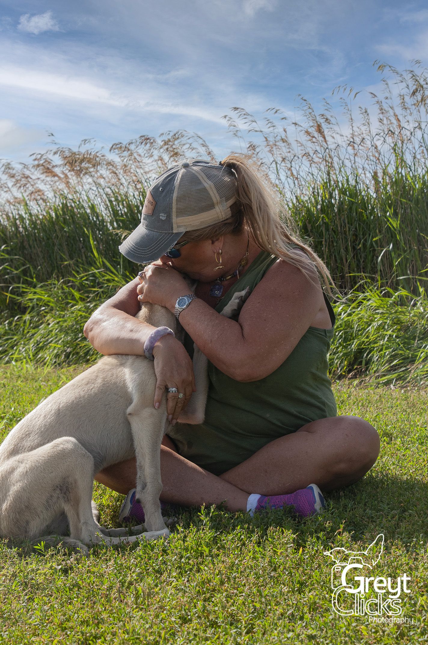 women wearing ballcap sitting in field hugging a fawn greyhound puppy