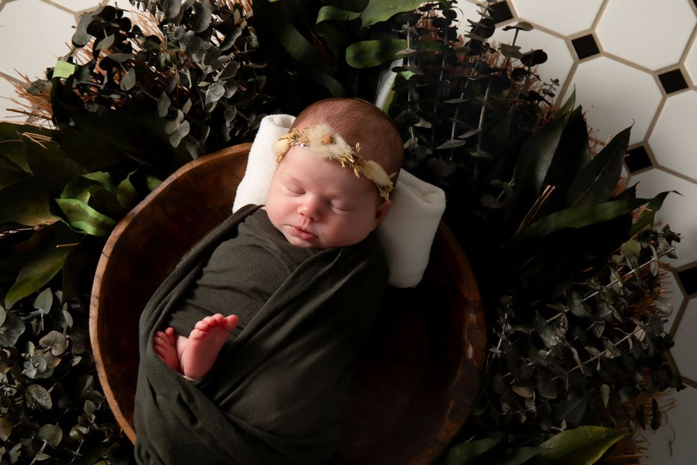 Sleeping newborn in dark wrap surrounded by greenery in wooden bowl.