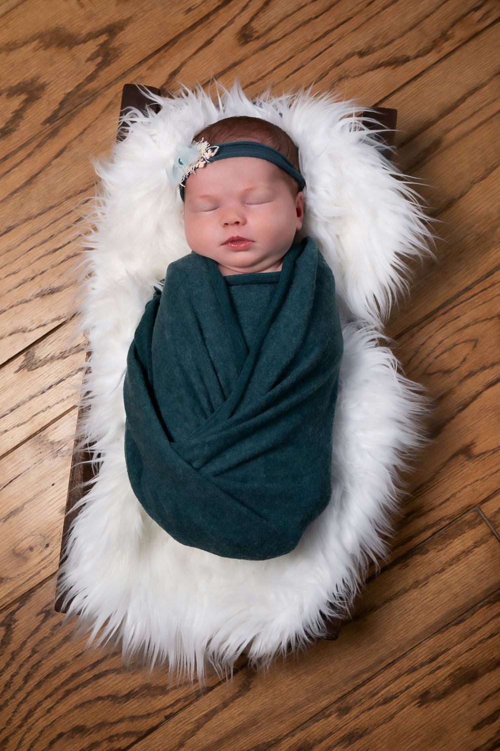 Peaceful newborn baby swaddled in teal fabric sleeping on white furry backdrop.