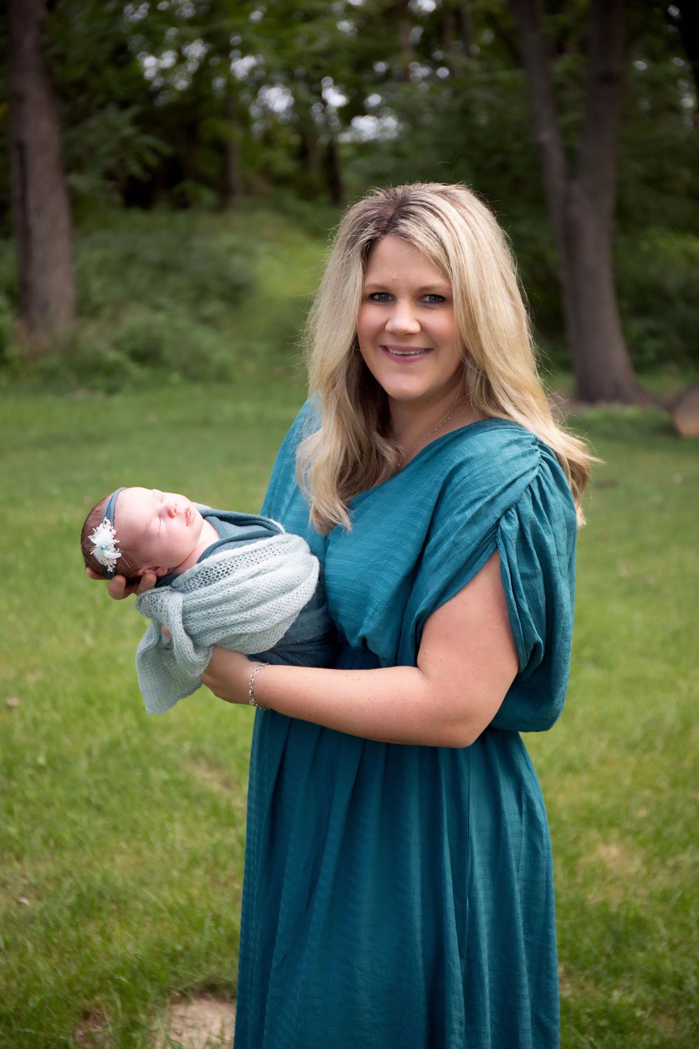 Woman in teal dress holds newborn baby outdoors in natural setting.