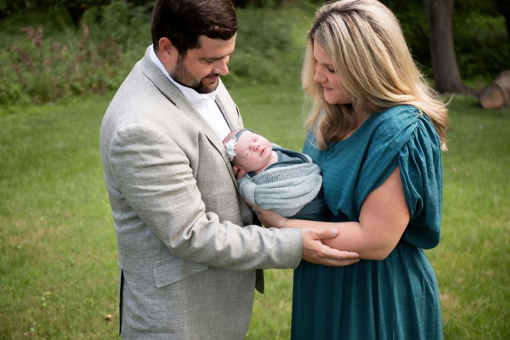 A couple holds their newborn baby outdoors in a grassy park setting.