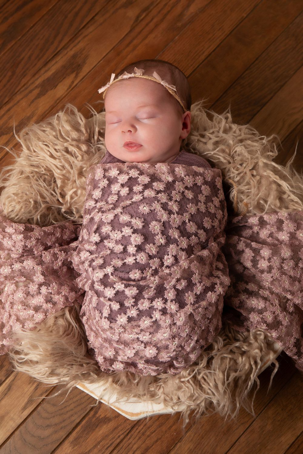 Sleeping baby wrapped in pink textured swaddle on cream fur against wooden floor.
