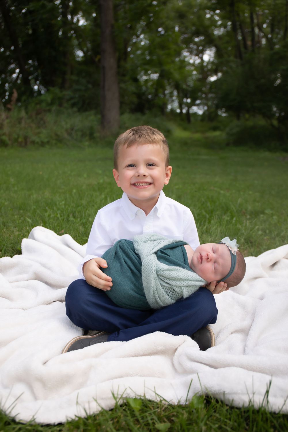 Two young children sit together on a white blanket in a grassy outdoor setting.