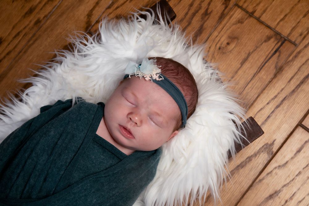 Sleeping newborn wrapped in teal swaddle on white fur rug against wooden floor.