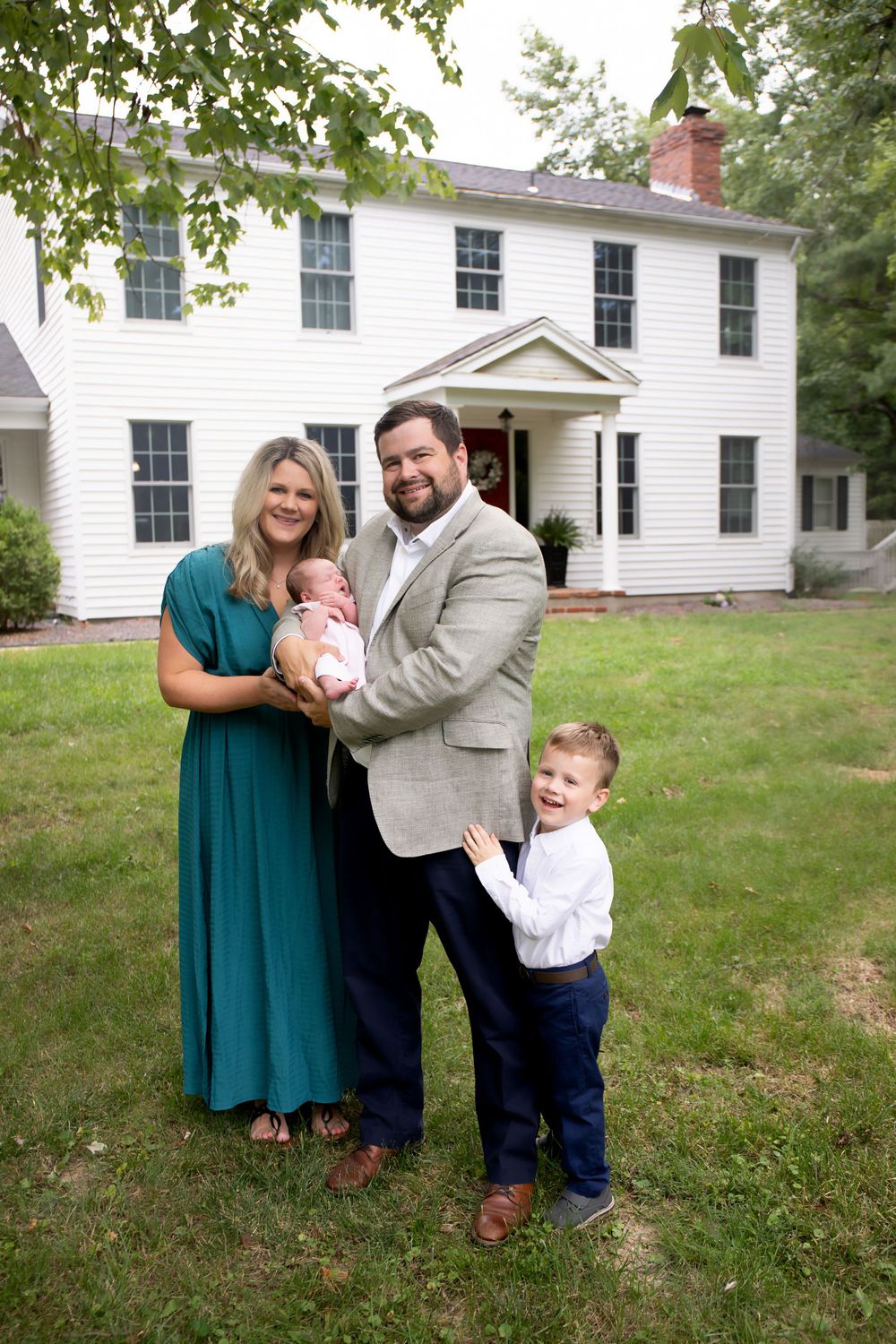 Family portrait in front of white colonial house with newborn baby and toddler.