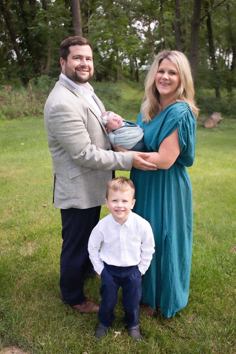 Outdoor family portrait with toddler and newborn baby against wooded background.