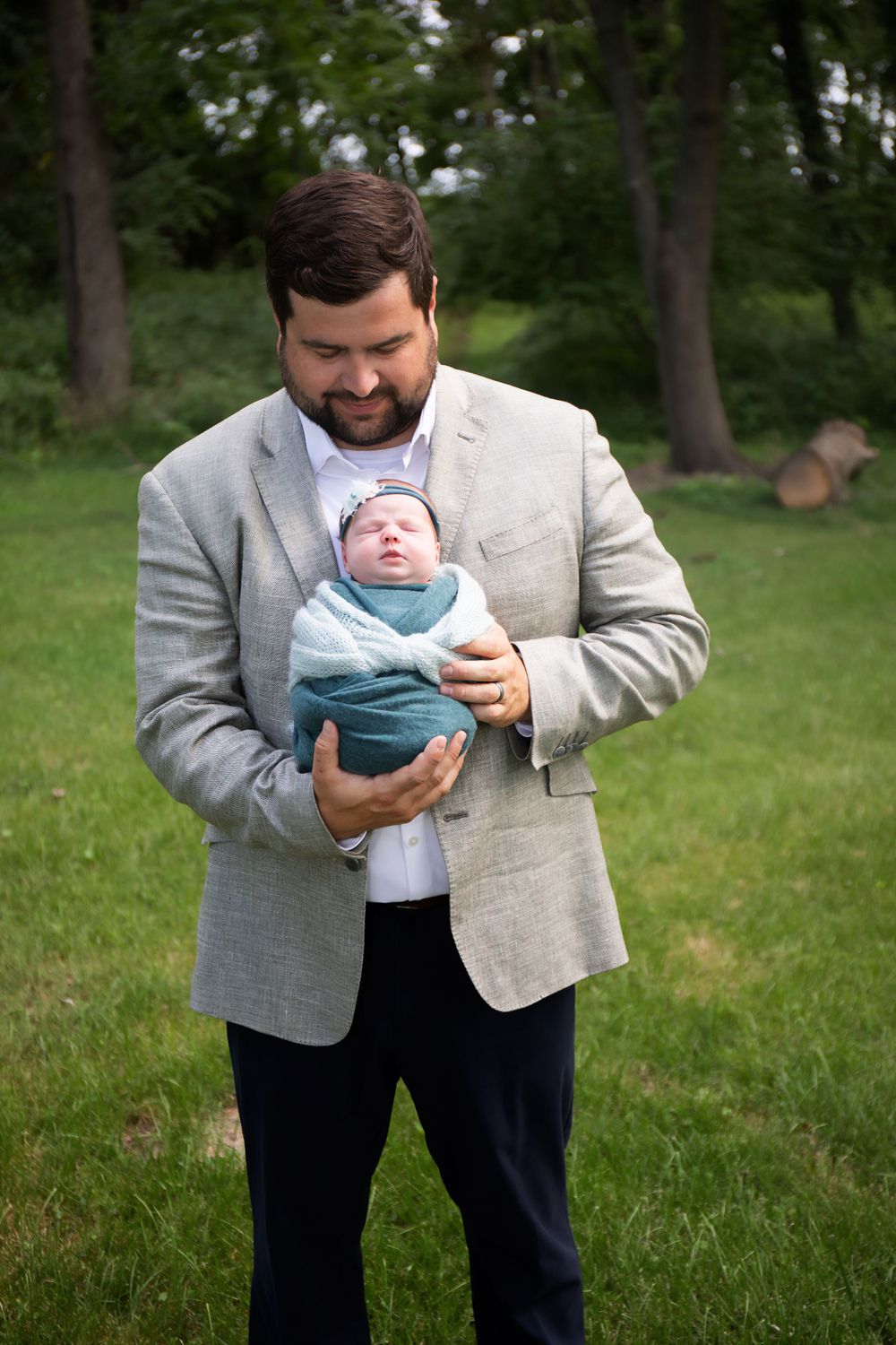 A person in a gray blazer cradles a swaddled newborn baby against a wooded background.