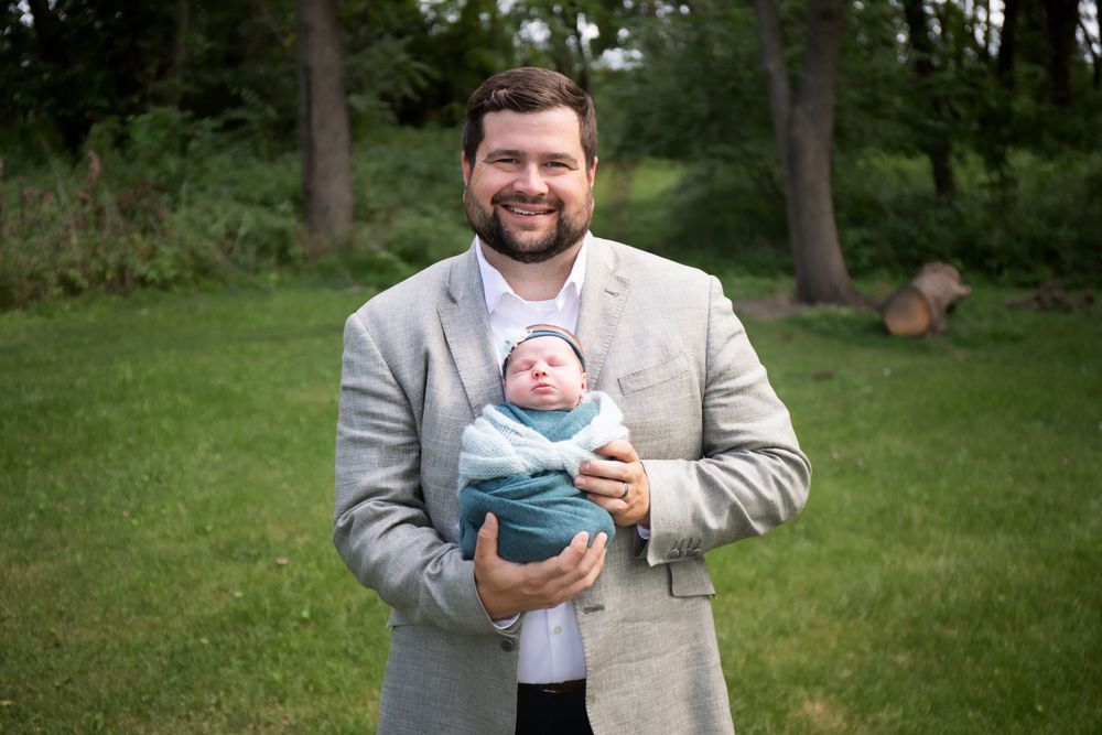 A beaming adult holds a tiny swaddled infant outdoors on a sunny day.