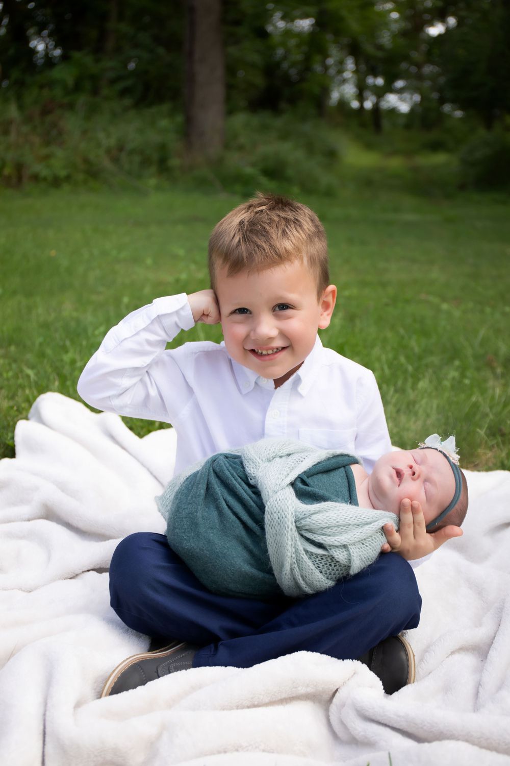 Two siblings pose on a white blanket outdoors, with the older one holding the baby.