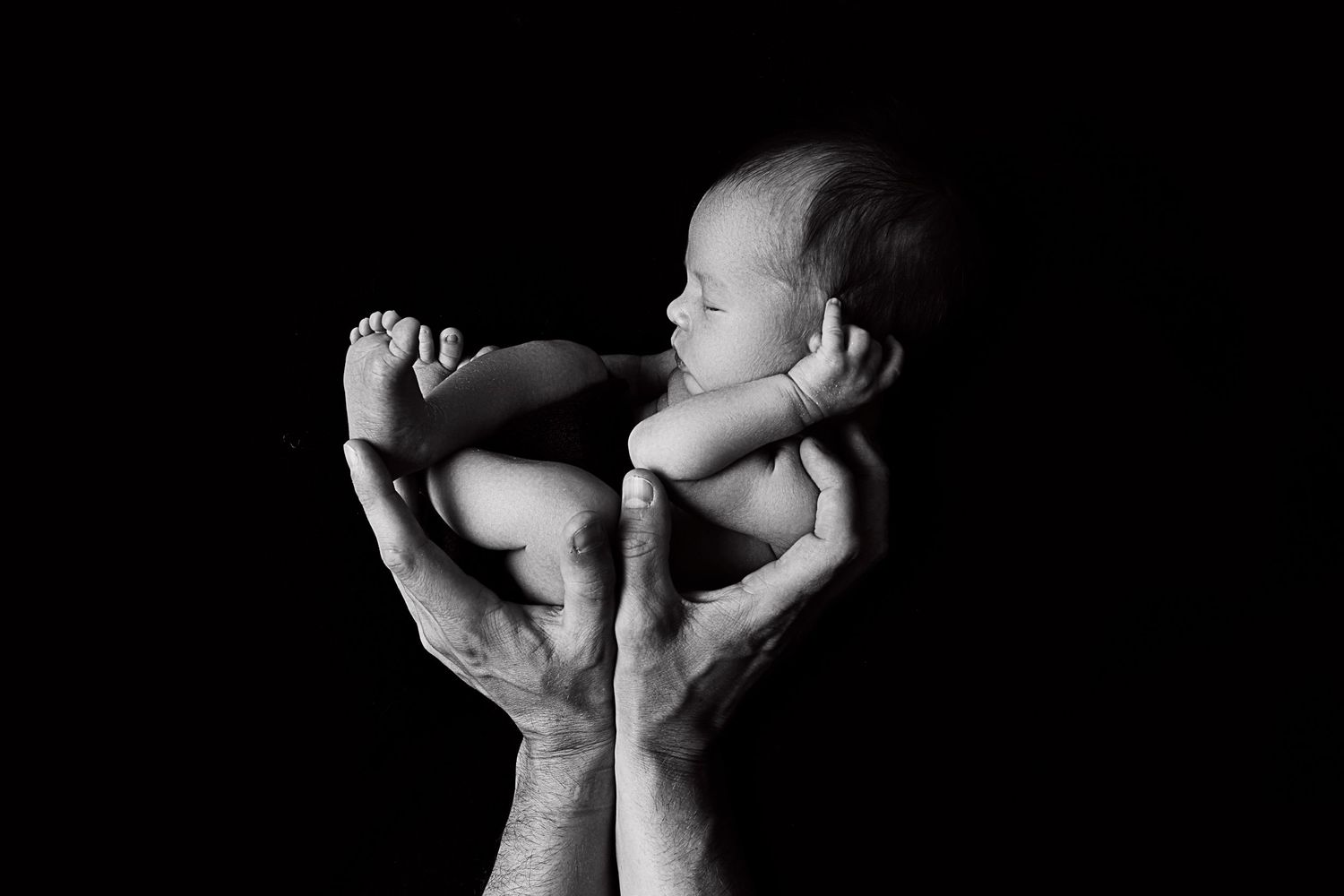 Gentle hands hold a peaceful newborn baby against dark backdrop.