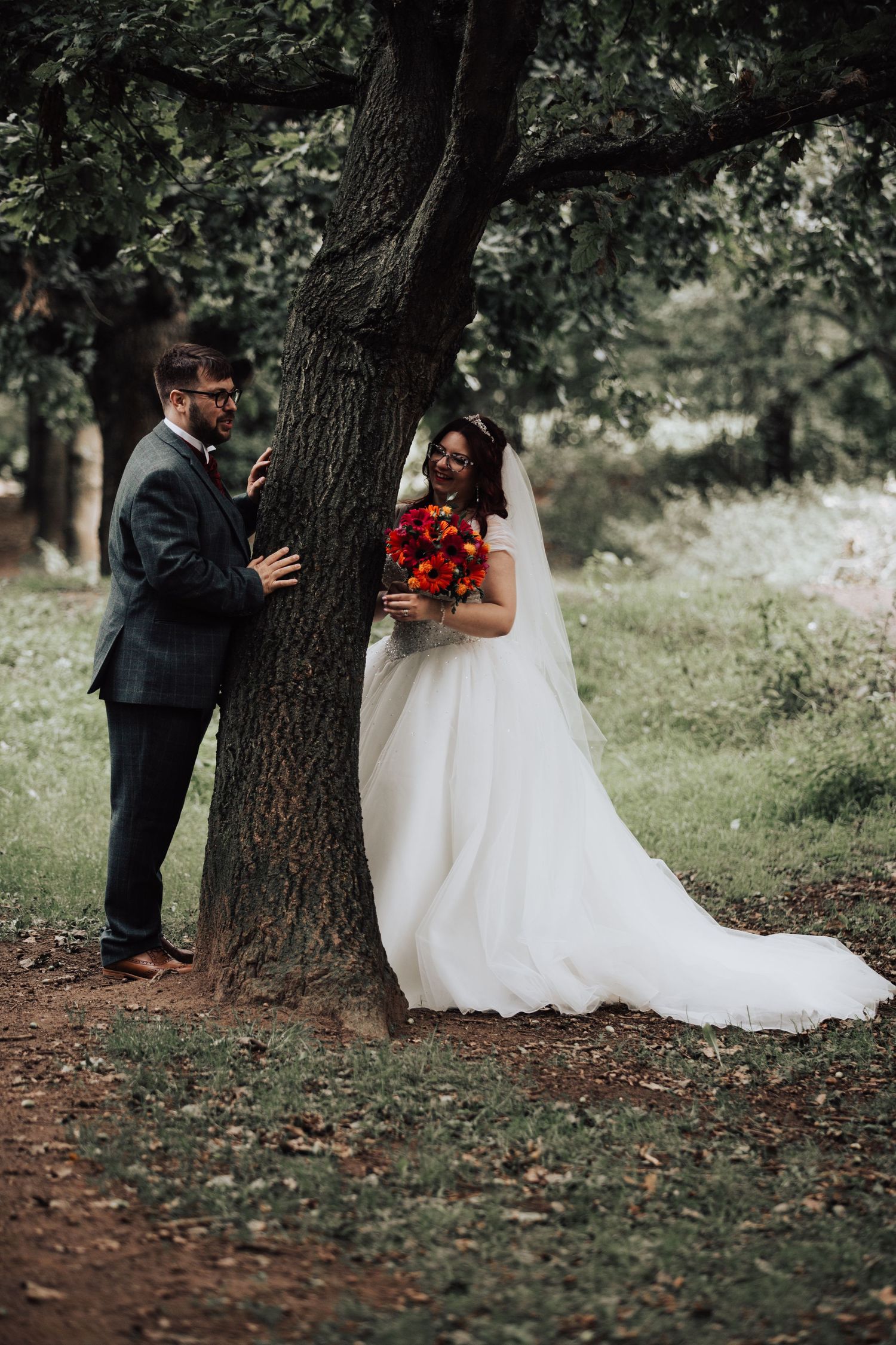 Wedding couple plays peek-a-boo around large tree trunk in natural outdoor setting.