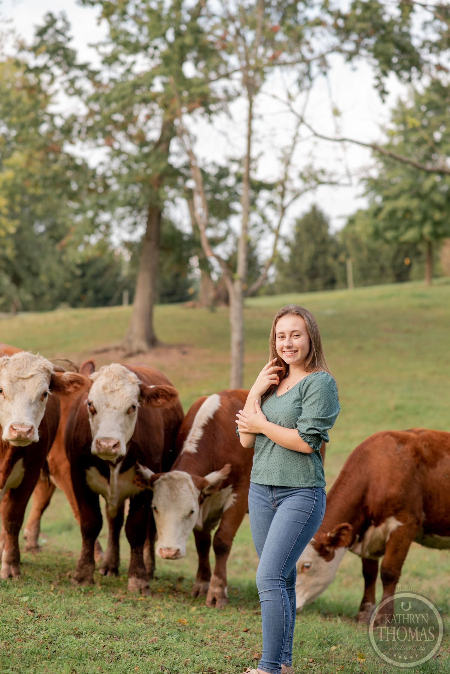 A person in a green shirt stands with a herd of Hereford cattle grazing on a grassy hillside.