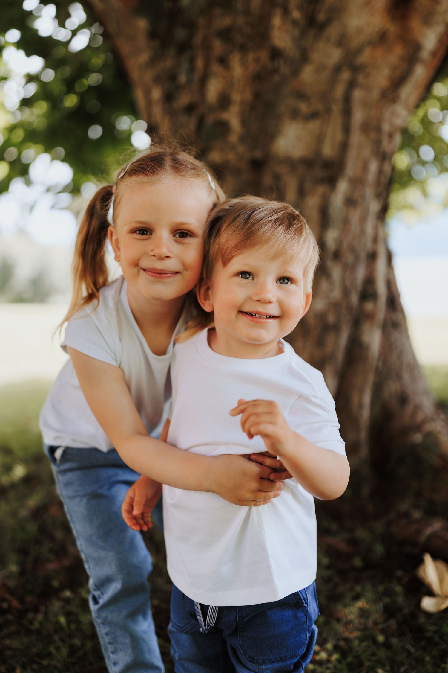 Two young children in white shirts and jeans hugging and smiling in front of a large tree trunk.