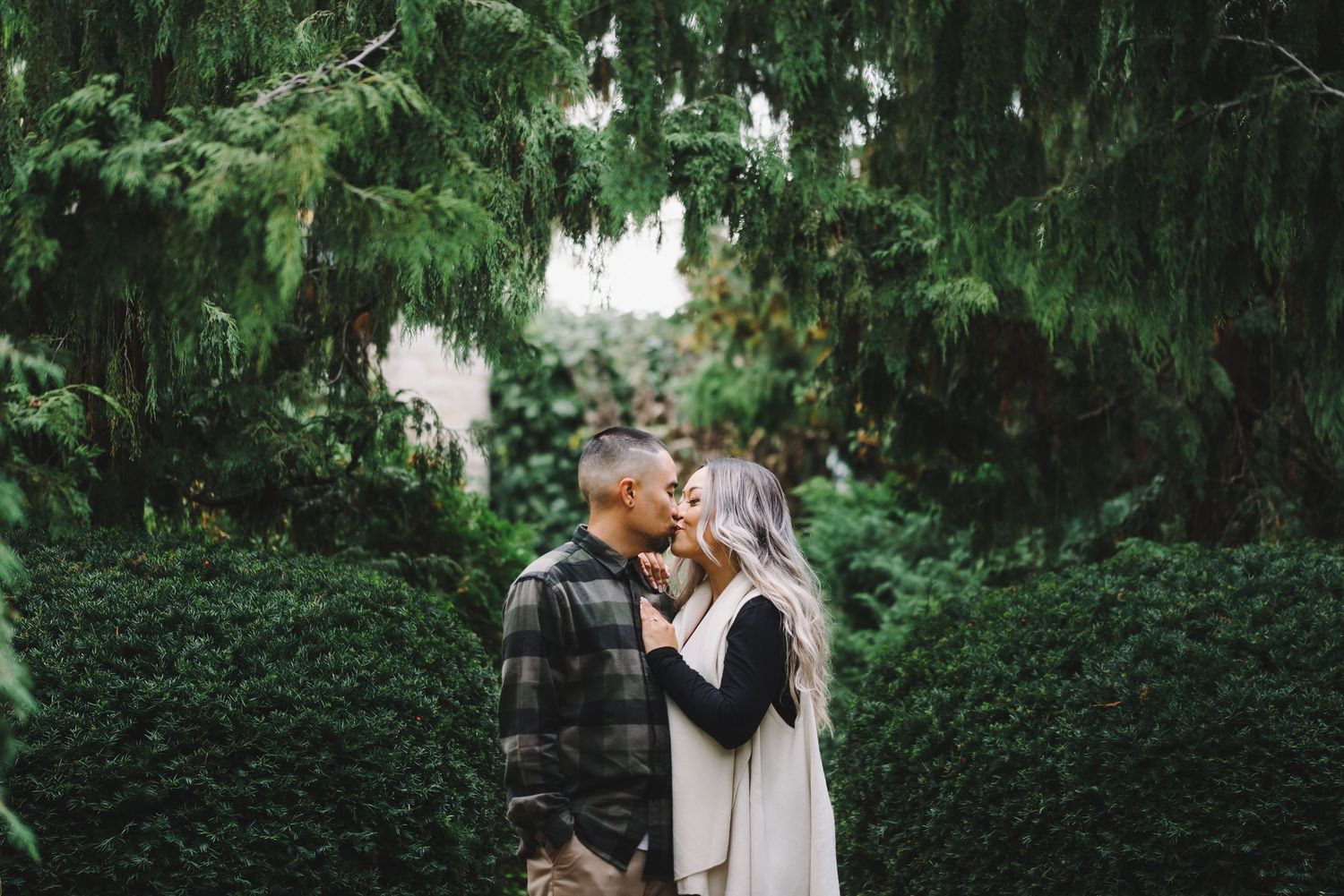 A tender romantic moment shared between hedges in a lush garden setting.