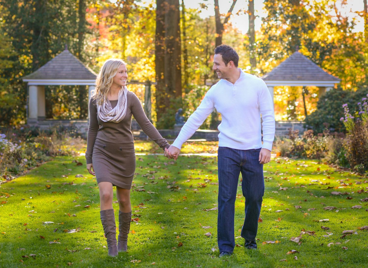 A couple holds hands while walking through a sunlit autumn garden with gazebos in the background.