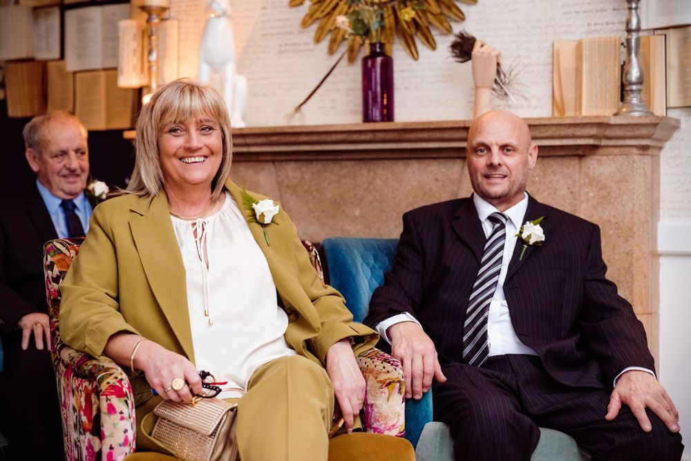 Wedding guests in formal attire seated on ornate furniture in front of a brick fireplace.