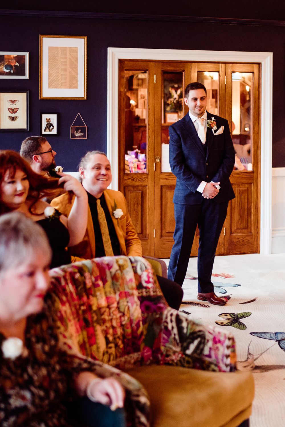 Wedding guests seated on floral furniture while someone in a navy suit stands near wooden doors.