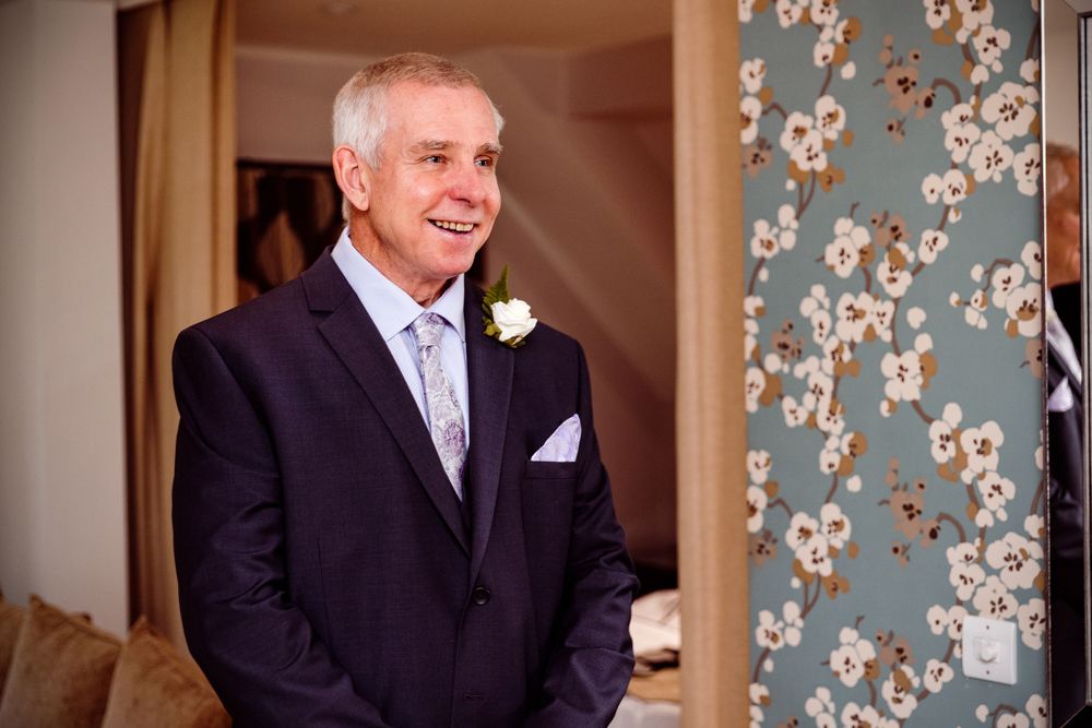 A person in a navy suit with a blue tie and white boutonniere smiling warmly by a floral patterned wall.