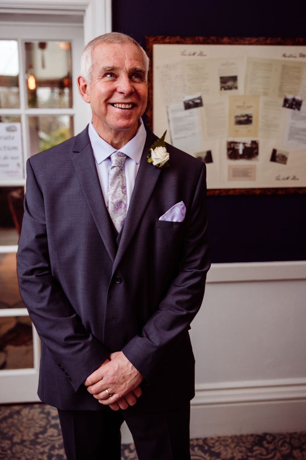 A person wearing a dark suit with lilac tie and white boutonniere smiling in an indoor setting.
