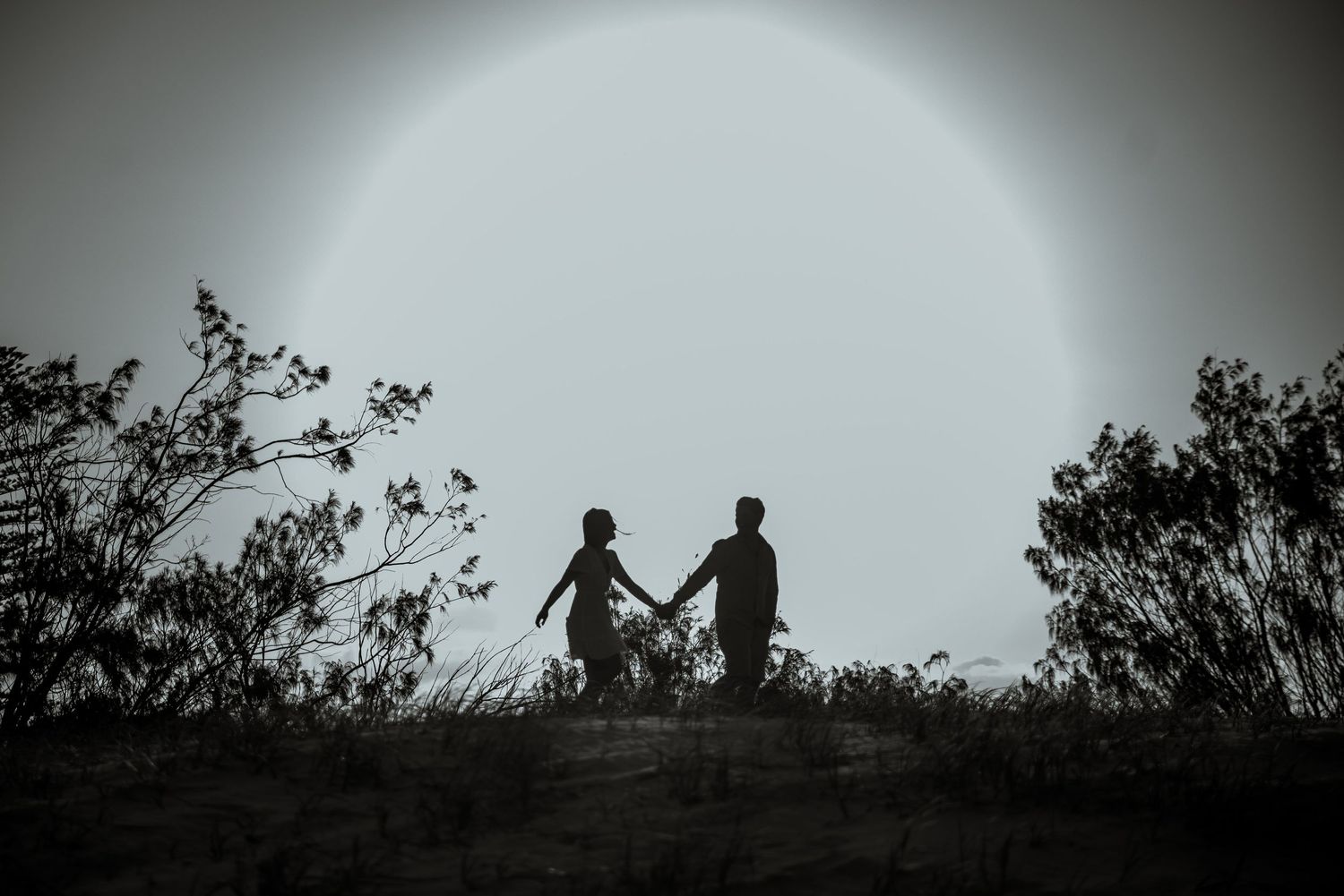 Silhouetted couple holding hands against a bright sunlit sky while standing on a hill.