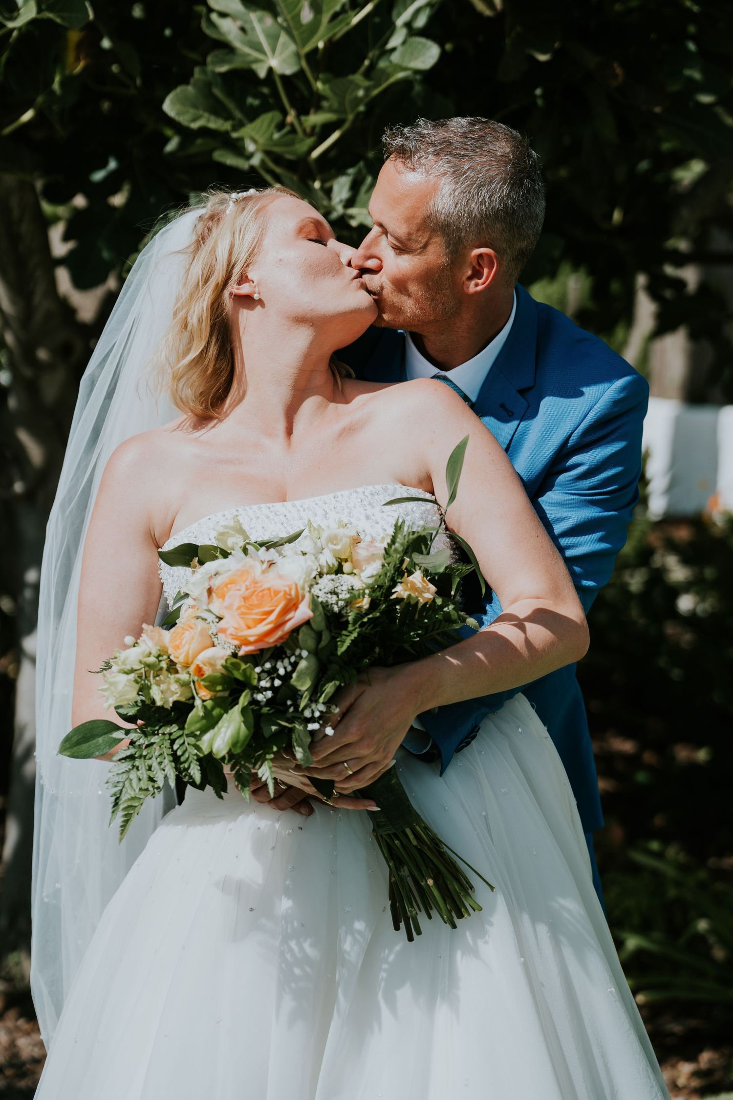 A romantic wedding photo of a couple kissing outdoors with the bride holding a peach and white floral bouquet.