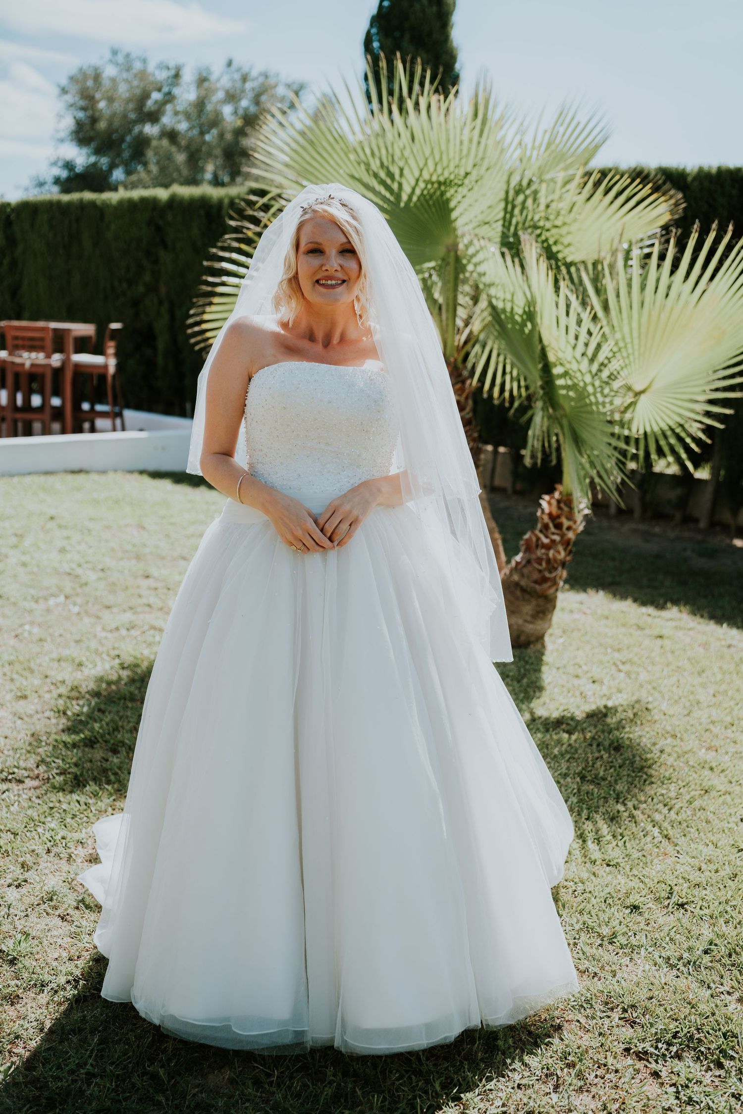 A bride in a flowing white ballgown and veil laughs joyfully in an outdoor garden setting.