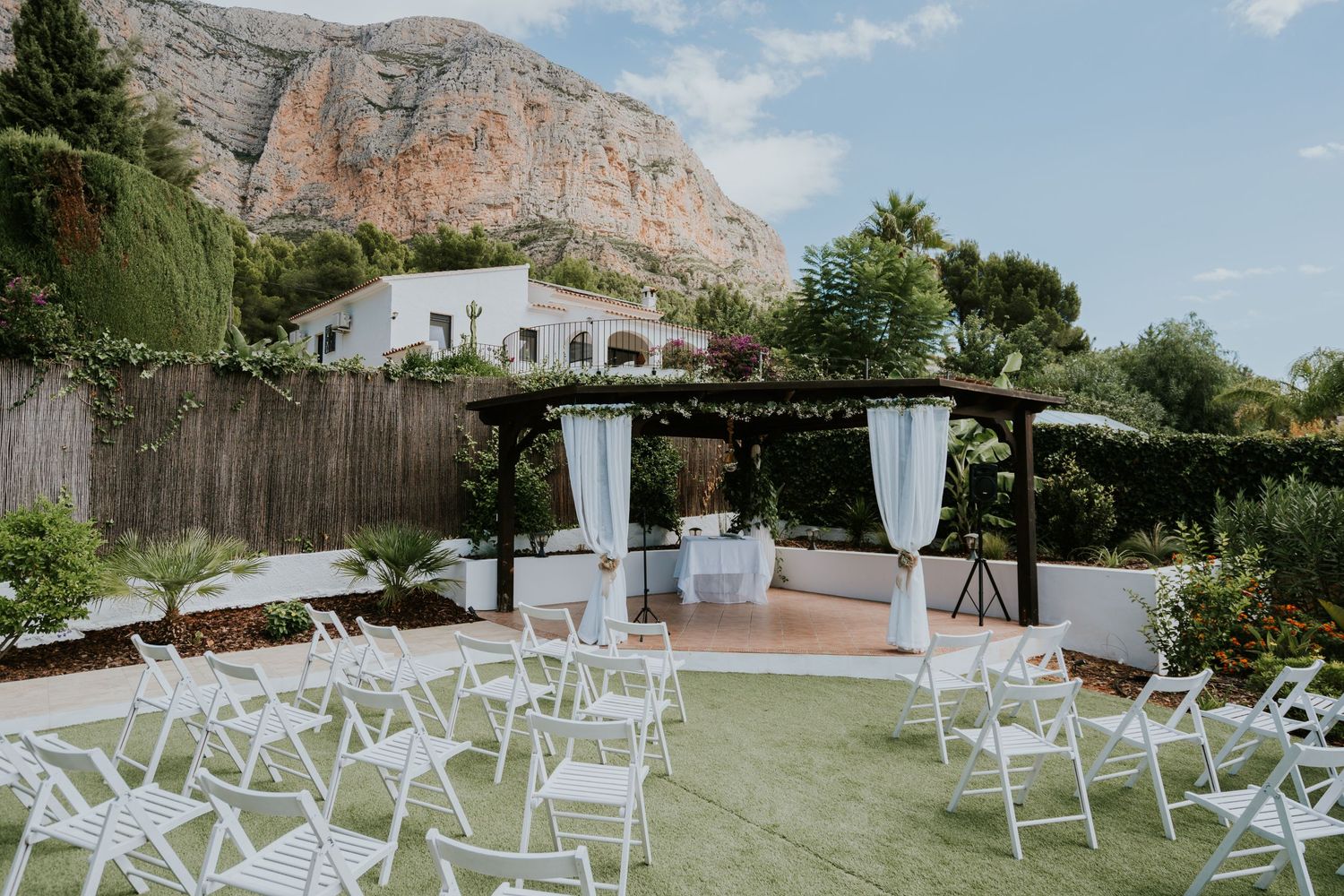 Outdoor wedding ceremony setup with white chairs and draped pergola against a mountain backdrop.