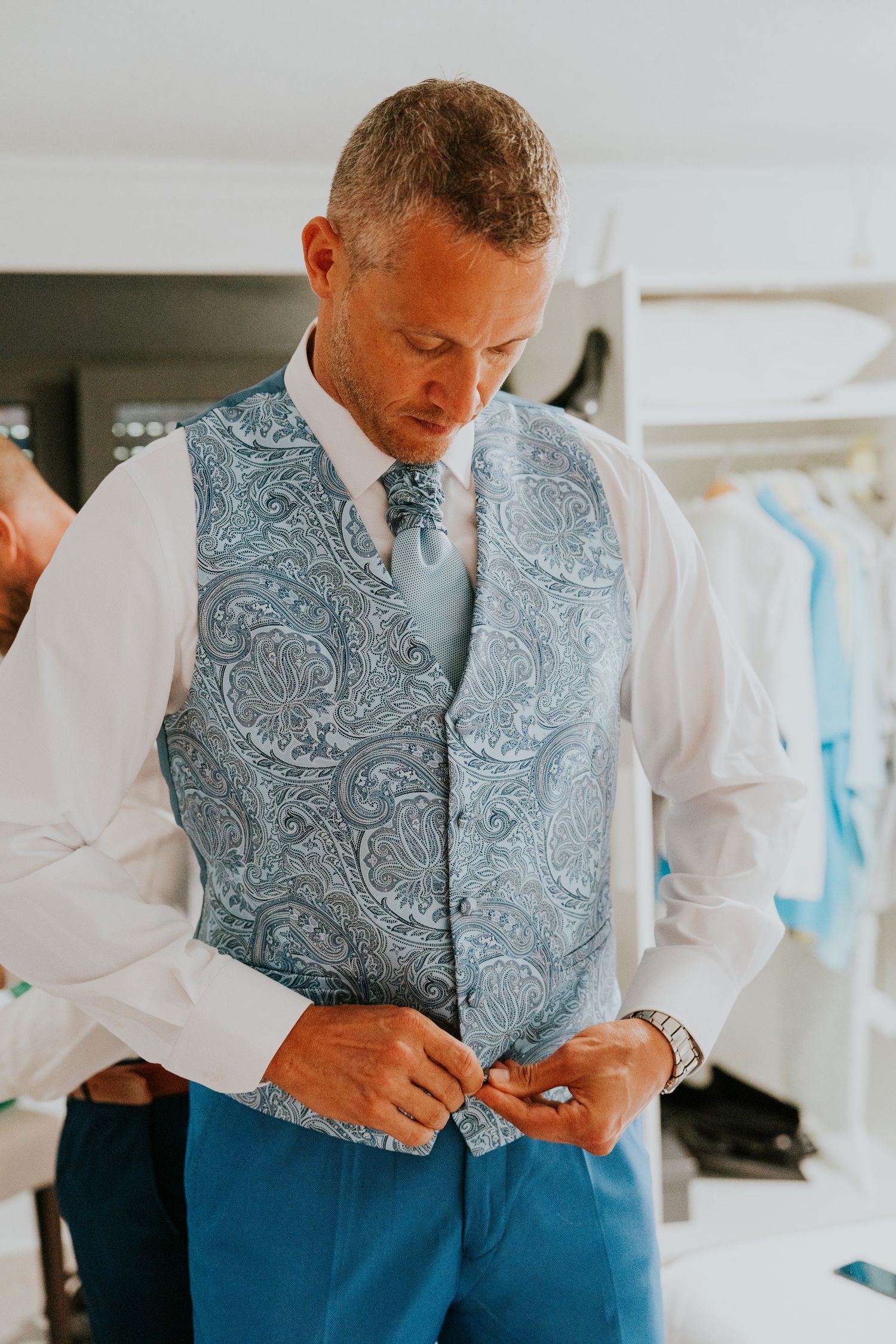 Someone adjusting formal attire wearing a light blue paisley vest and white dress shirt.