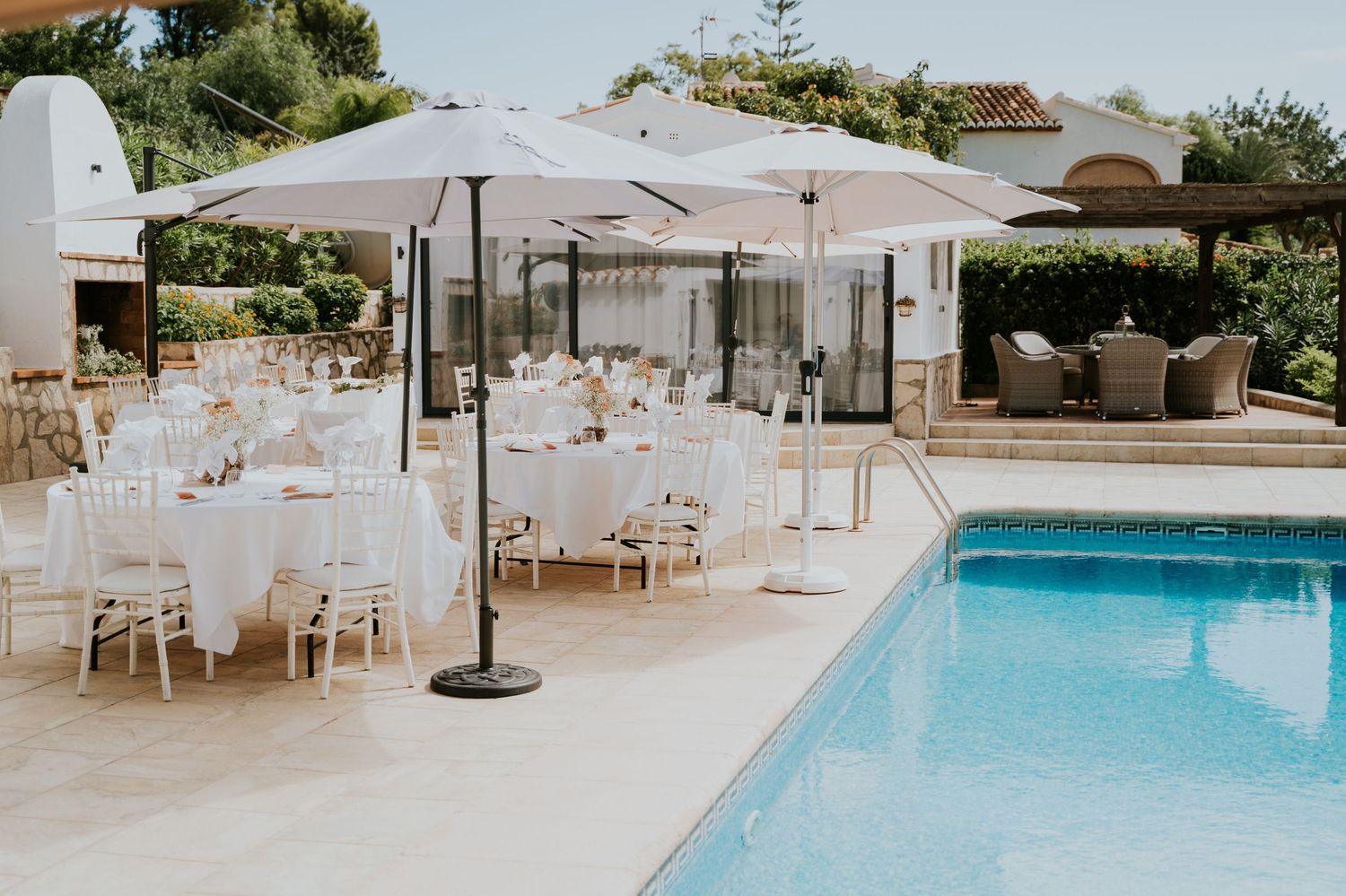Elegant outdoor wedding reception setup with white tables and chairs around a swimming pool.