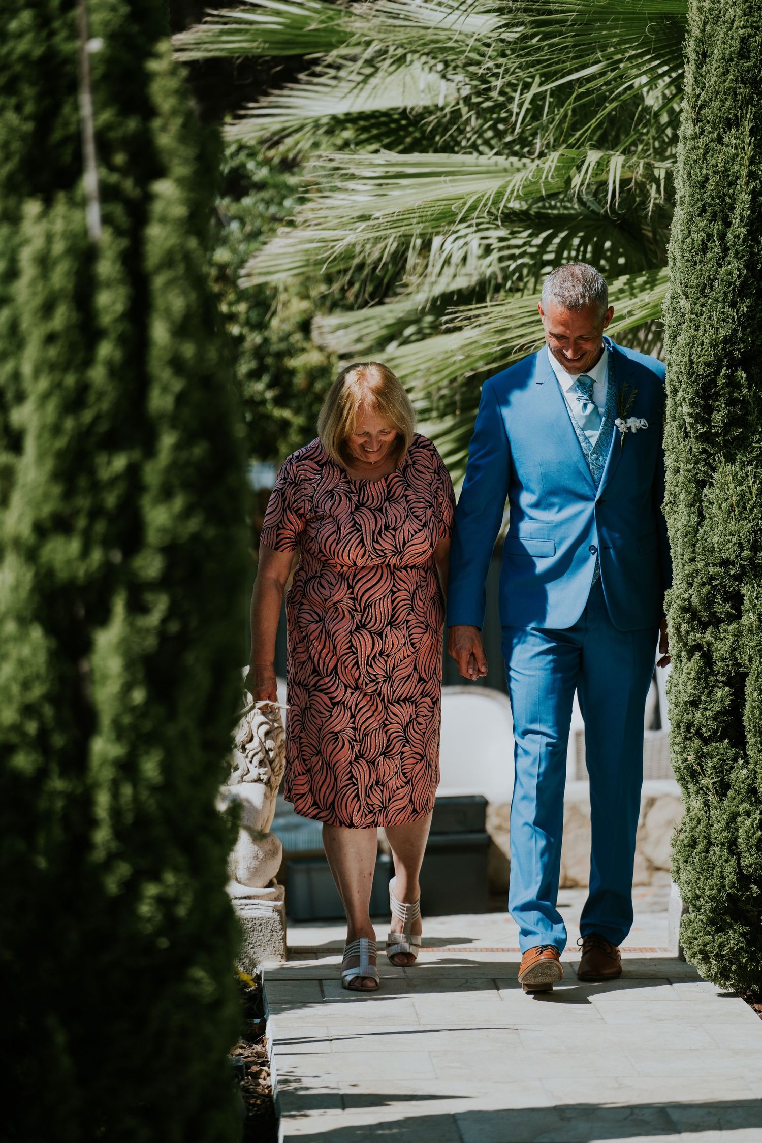 Two people walking down a palm-lined pathway, one in blue suit and one in patterned dress.
