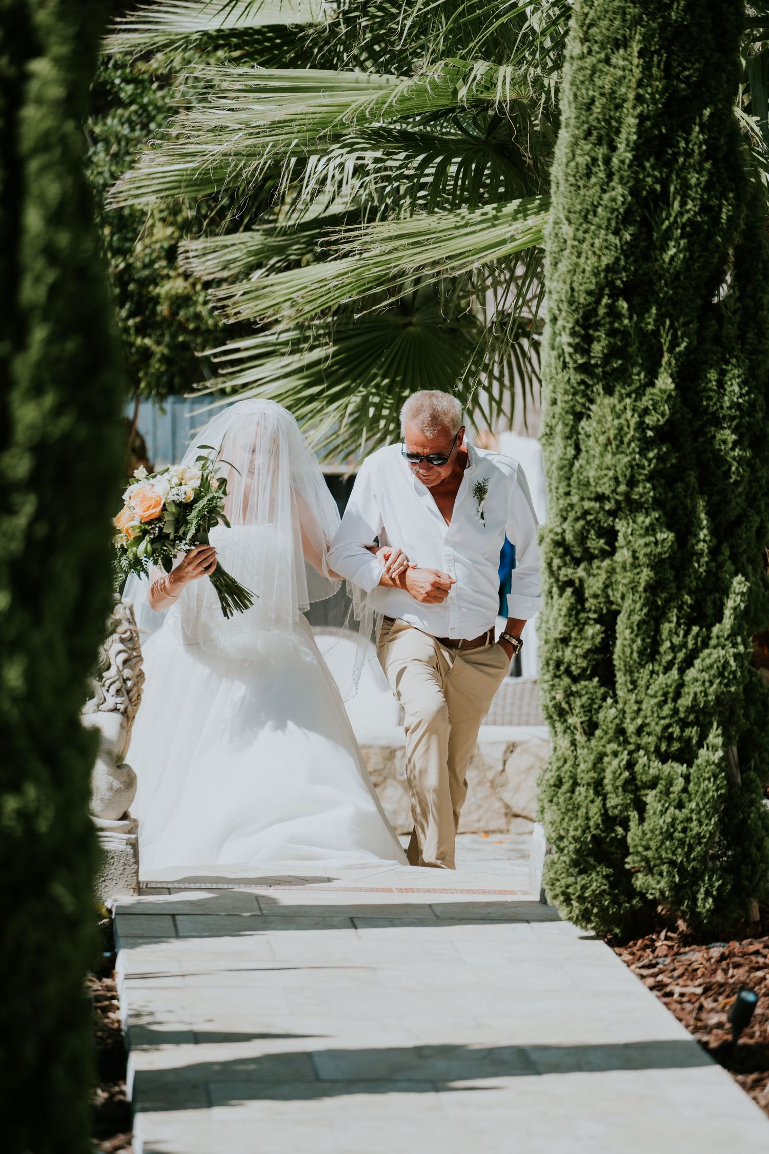 A wedding ceremony walkway lined with tall cypress trees and palm fronds as a couple walks together.