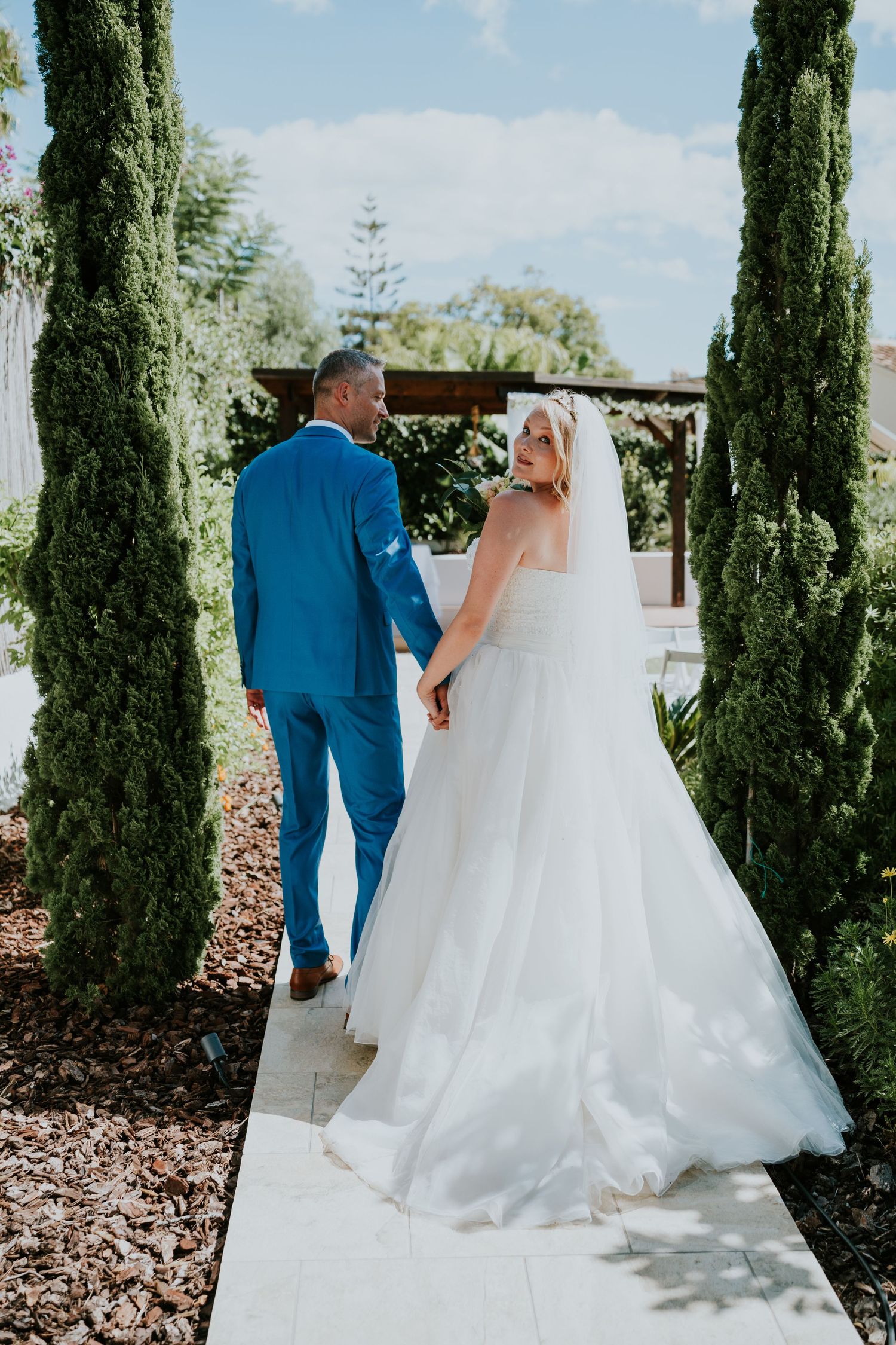 A bride and groom walking along a cypress-lined pathway in their wedding attire.