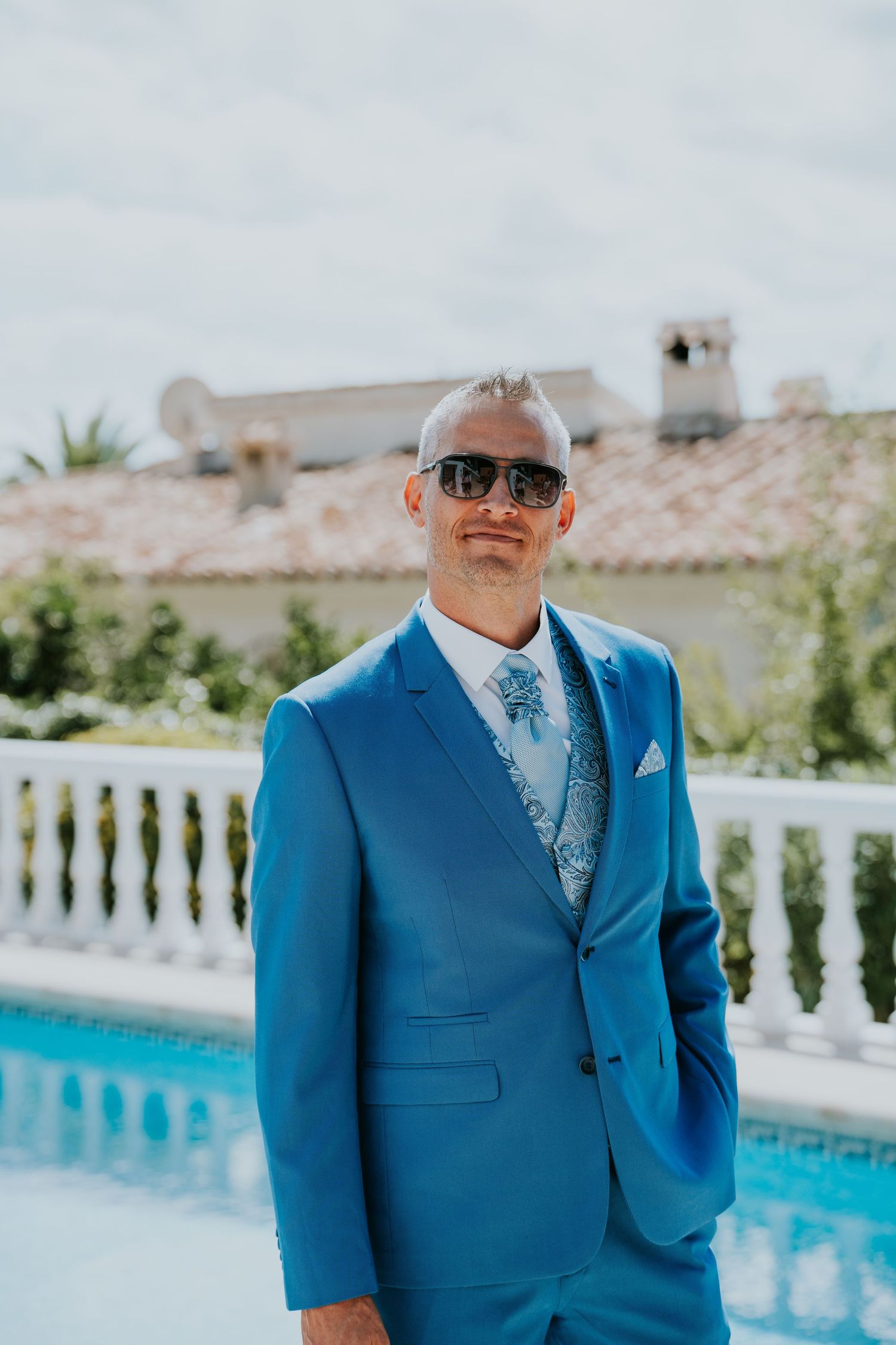 A formal portrait of someone in a bright blue suit with matching paisley vest by a pool.
