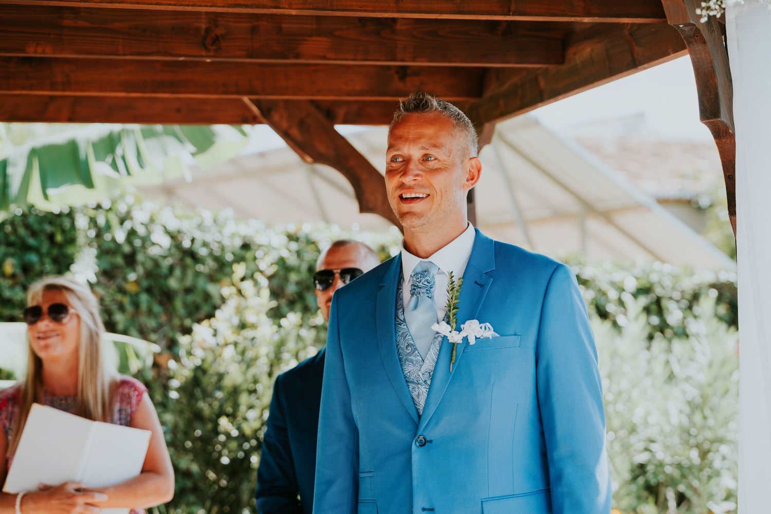 Someone in a bright blue suit standing under wooden beams at an outdoor ceremony.