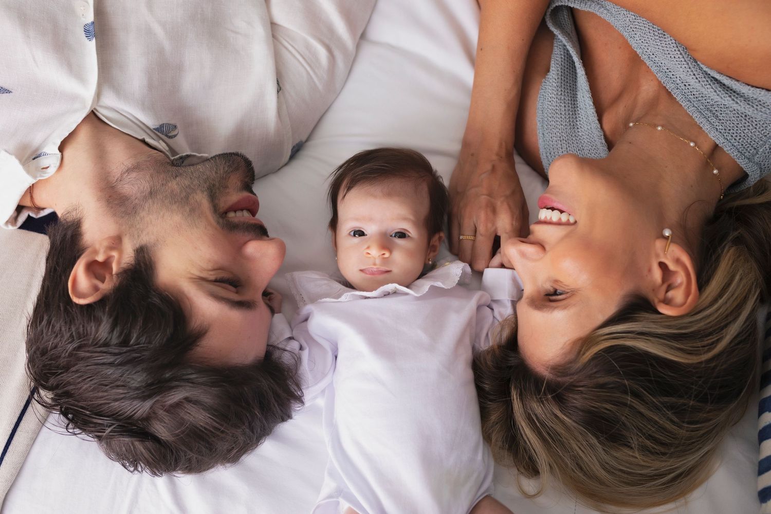 A baby wearing white lies between two adults on light bedding.