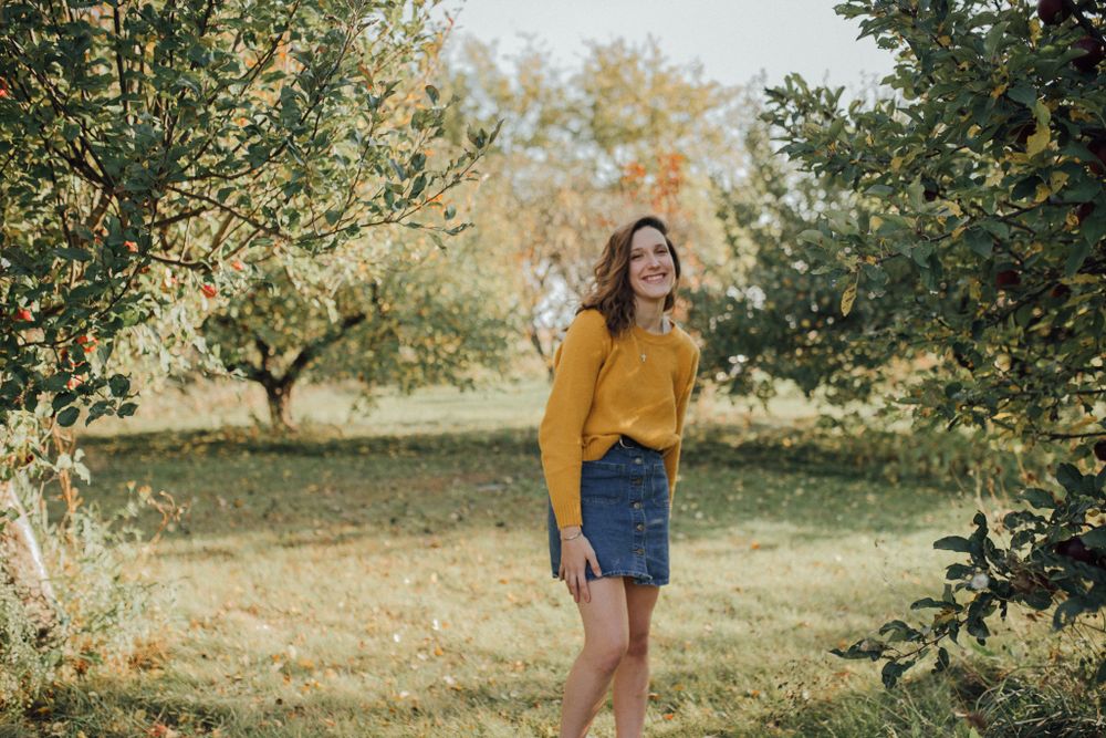 Person in yellow sweater and denim skirt standing among autumn orchard trees.